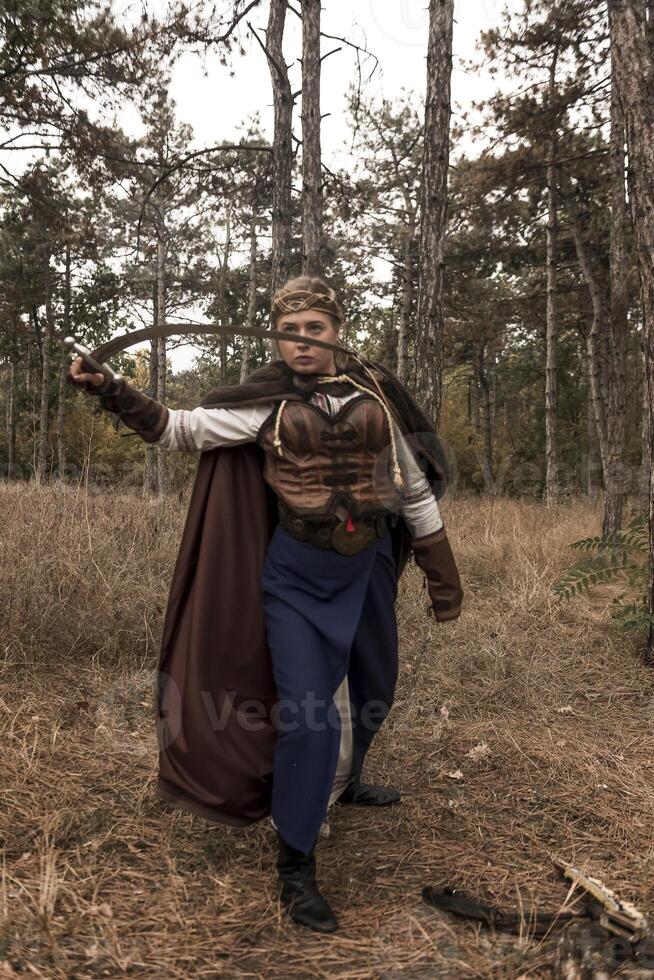 warrior girl with sword in hands in forest. blonde dressed in historical costume of warrior woman, with long cloak. warrior girl, raised sword, attacks enemy, looks at camera photo