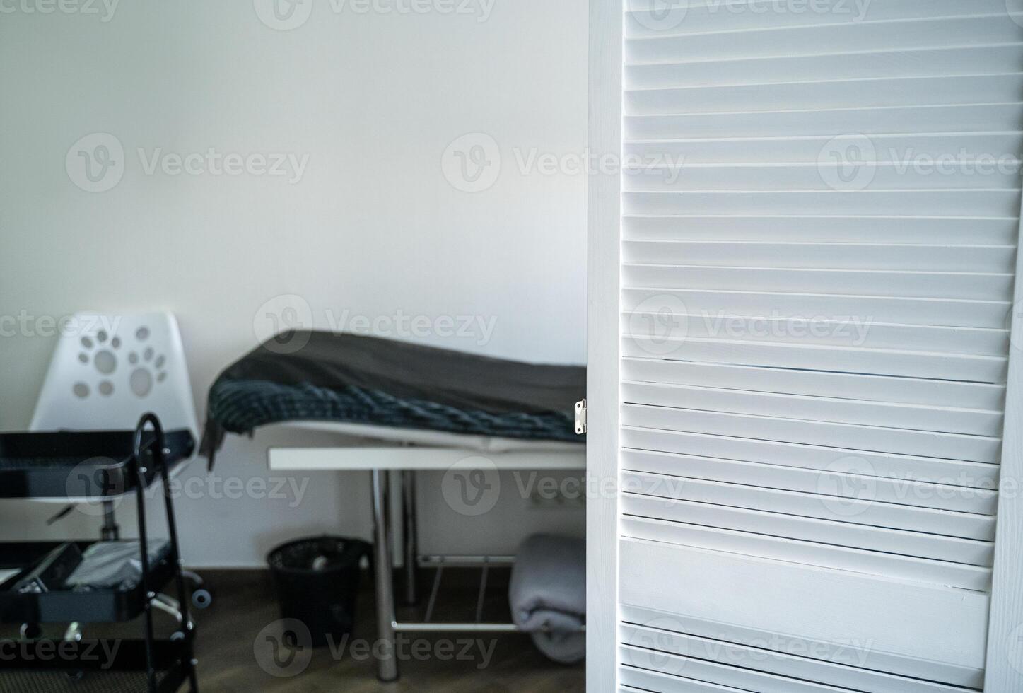 table for massage and other cosmetology services. covered with a blanket and a disposable sheet. next to it is a white chair. in the foreground is a white screen. photo