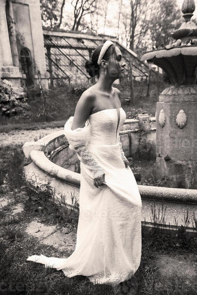 Bride with smooth hairstyle and white headband in hair against old building background. Brunette girl in long white dress with open shoulders. Ruins of 18th century fountain behind. photo