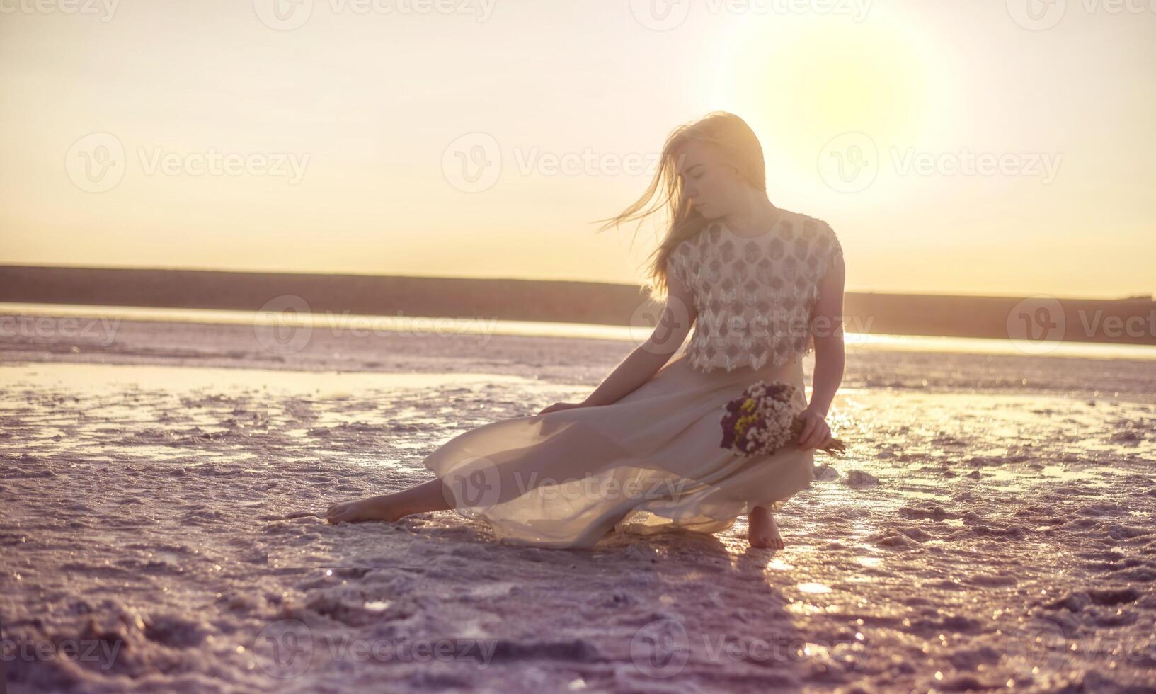 blonde girl sits in the middle of a salt lake. her dress flutters in the wind. a bright circle of the setting sun illuminates the shore covered with salt. she holds a bouquet in her hands, odessa photo