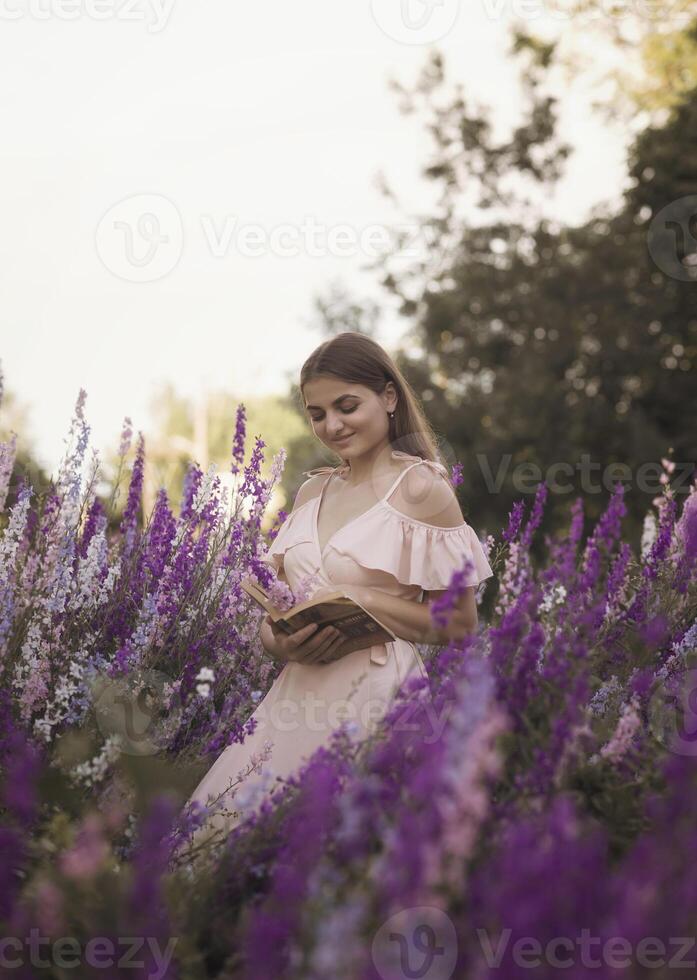 A beautiful girl stands in nature in the middle of a blooming field. She holds a book in her hands. There are pink and purple flowers around her. Her dress is soft pink. Behind are trees. photo