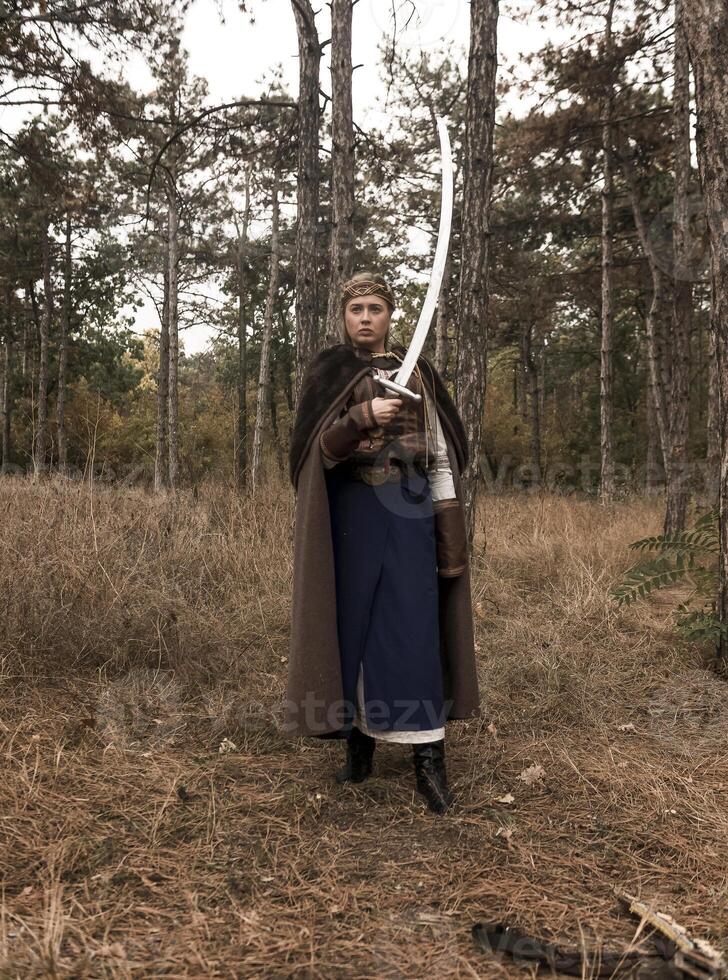 warrior girl with sword in hands in forest. blonde dressed in historical costume of warrior woman, with long brown fur cloak and medieval dress. warrior girl holds steel sword in hands, front view photo