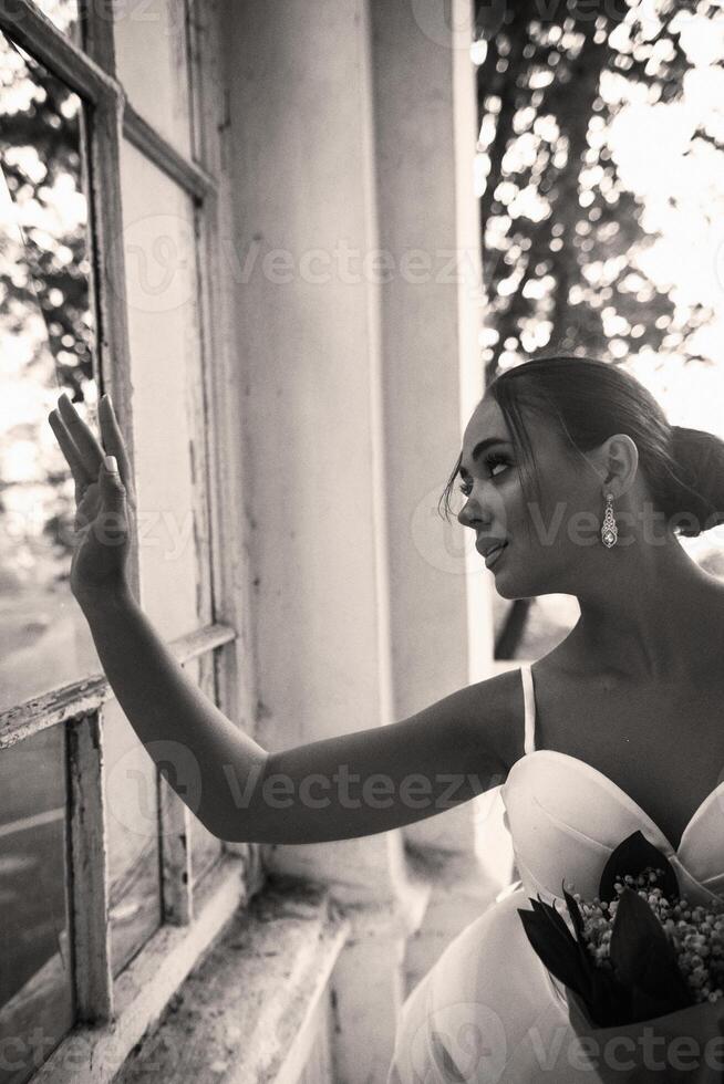 a beautiful girl in a white dress with a bouquet of lilies of the valley stands near the window. the brunette has an elegant hairstyle, styled in a bun. black and white photo, vertical orientation. photo