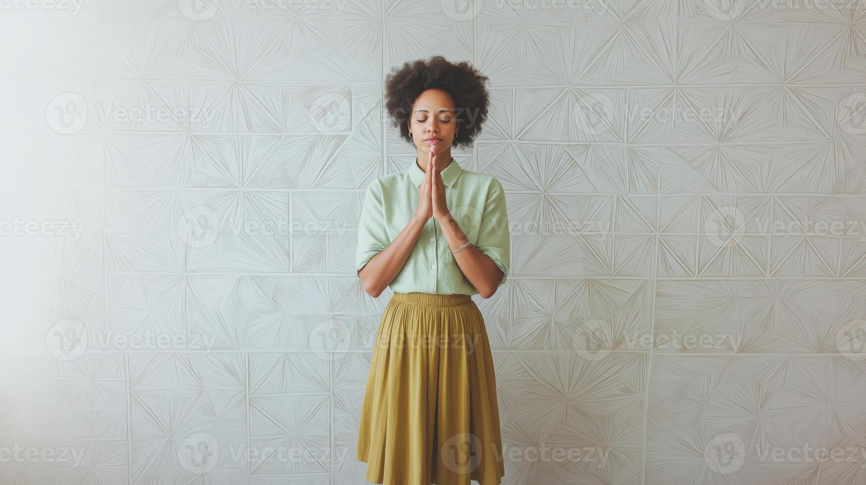 woman standing in front of a white wall with a geometric pattern. She is wearing a light green button-down shirt and a mustard-colored skirt. photo