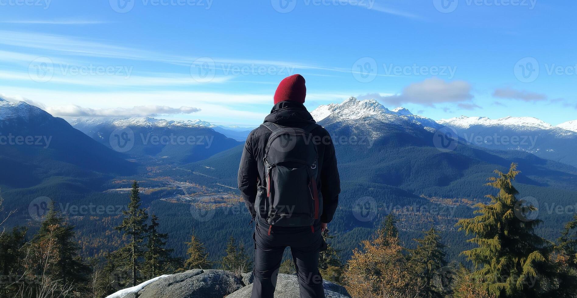 man standing on top of a mountain, looking out at a beautiful landscape. He is wearing a black jacket, a red beanie, and carrying a large black backpack photo