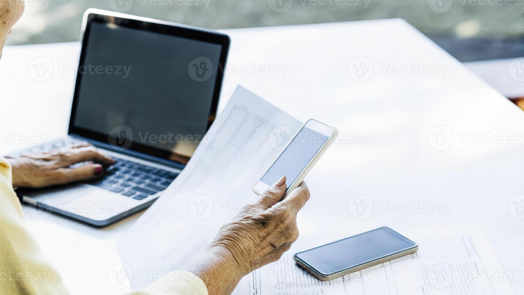An elderly womans hand sorting through printed health documents and notes surrounded by modern technology illustrating the blend of traditional and digital approaches to managing. photo