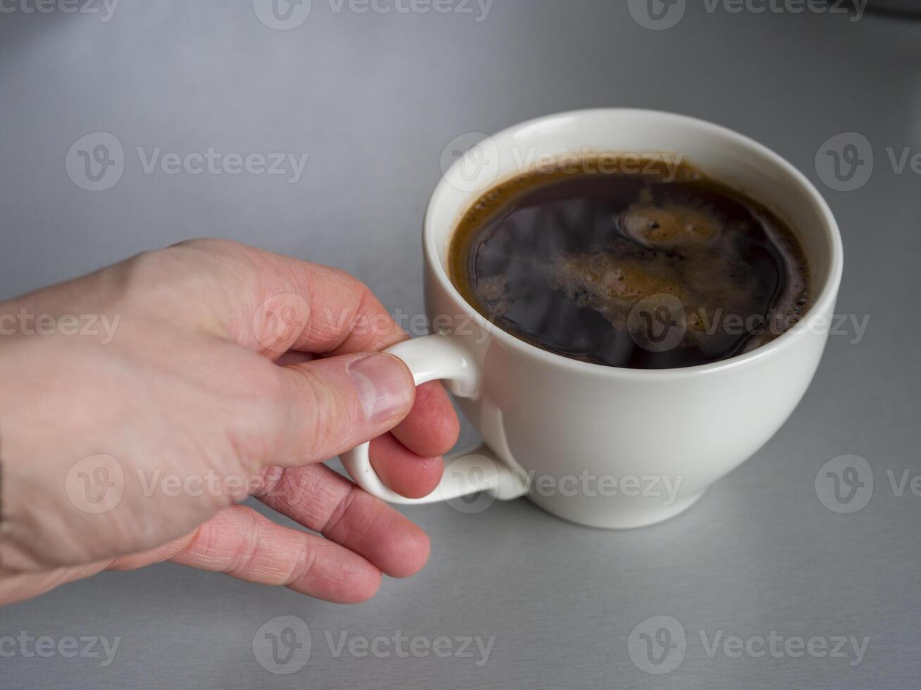 Hand gently grasps the handle of a white mug filled with dark, aromatic coffee, resting on a simple gray table, creating a cozy and inviting scene photo