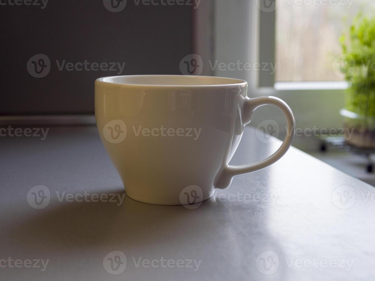 Large white coffee mug resting on a kitchen counter, positioned in front of a window. Out-of-focus green plant on the windowsill adds a touch of nature to the serene, blurred background photo