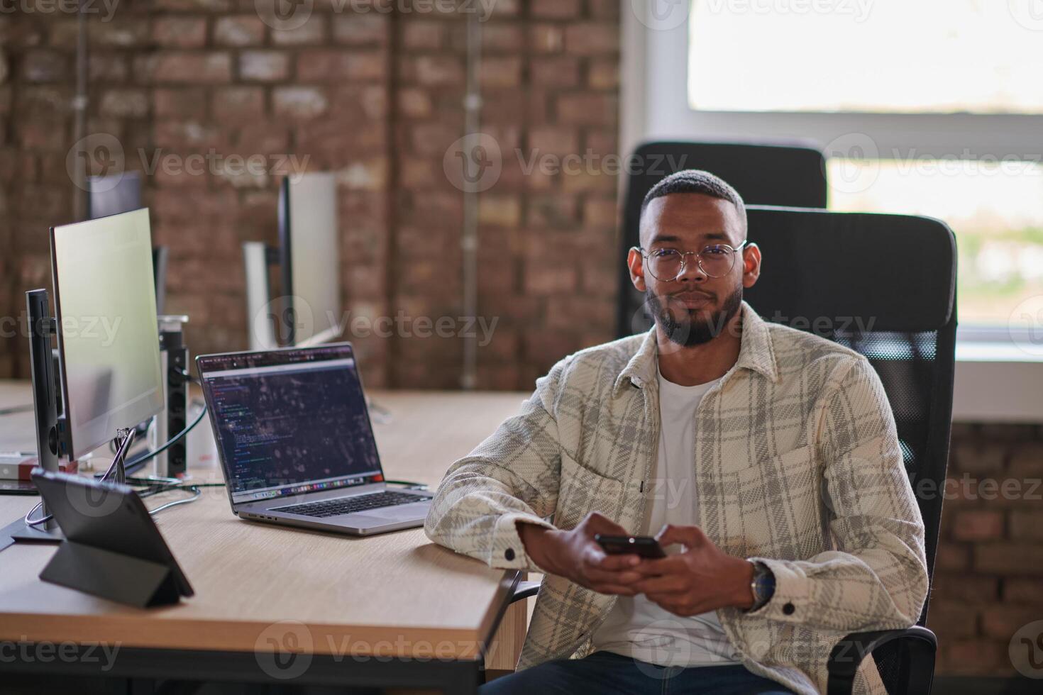 A young African American entrepreneur takes a break in a modern office, using a smartphone to browse social media, capturing a moment of digital connectivity and relaxation amidst his business photo