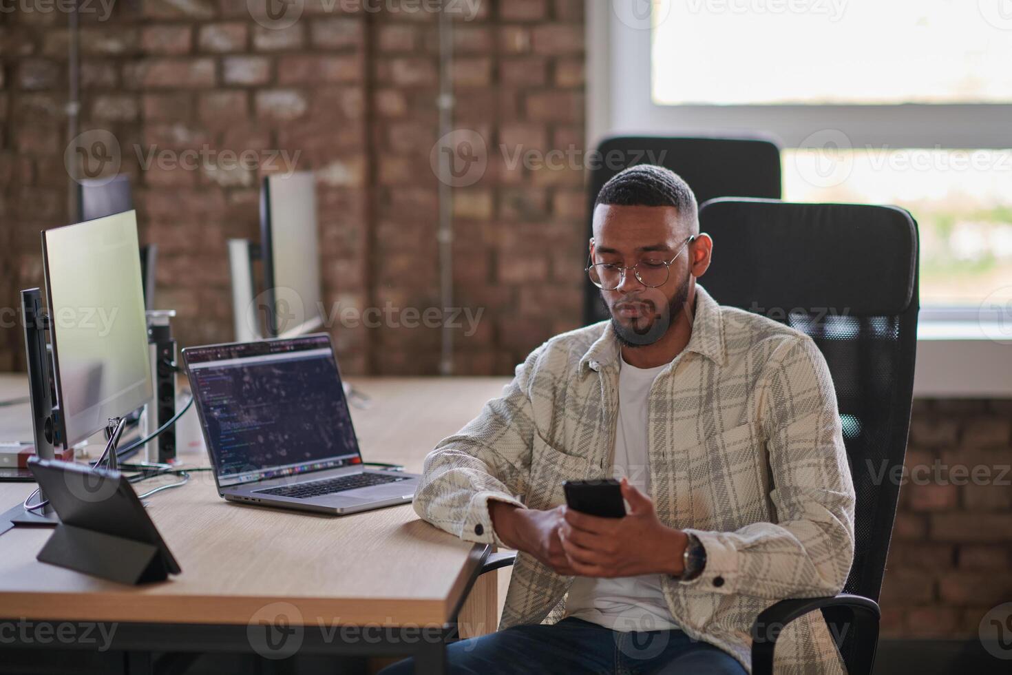 A young African American entrepreneur takes a break in a modern office, using a smartphone to browse social media, capturing a moment of digital connectivity and relaxation amidst his business photo