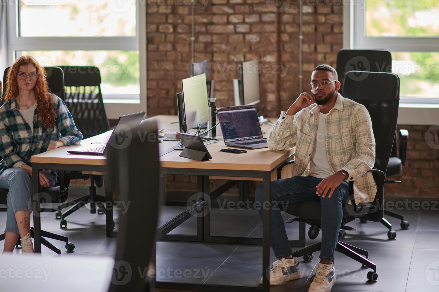 Group of colleagues, a woman with vibrant orange hair and a young African American businessman, sitting in a modern office space, symbolizing diverse collaboration and a dynamic work environment. photo