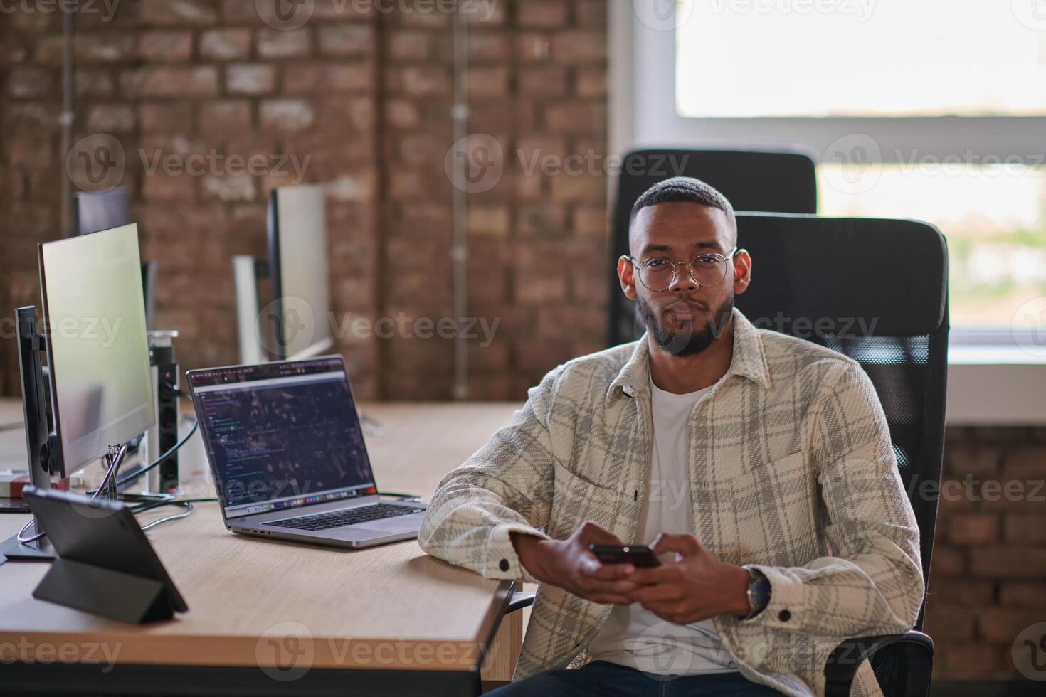 A young African American entrepreneur takes a break in a modern office, using a smartphone to browse social media, capturing a moment of digital connectivity and relaxation amidst his business photo