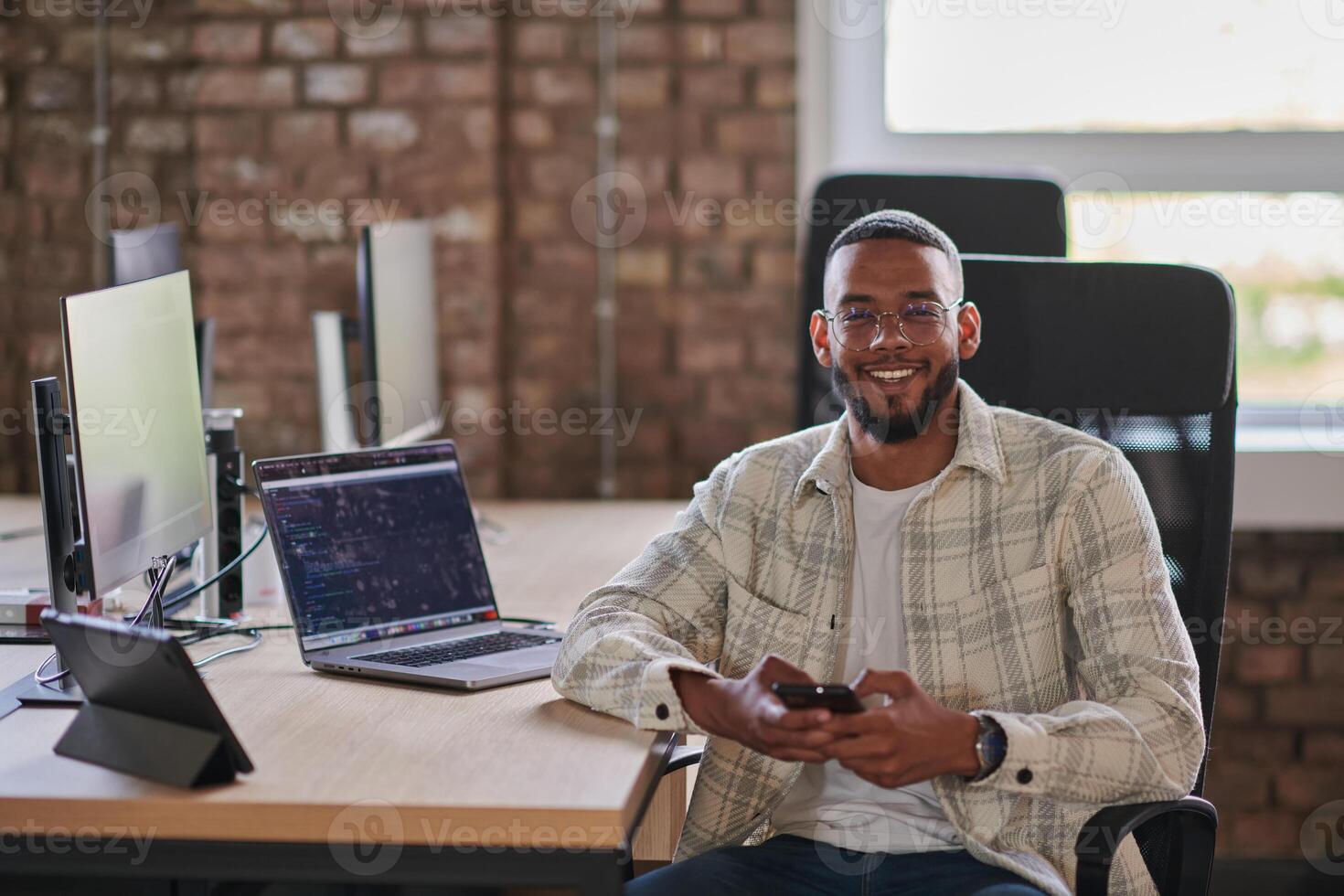 A young African American entrepreneur takes a break in a modern office, using a smartphone to browse social media, capturing a moment of digital connectivity and relaxation amidst his business photo