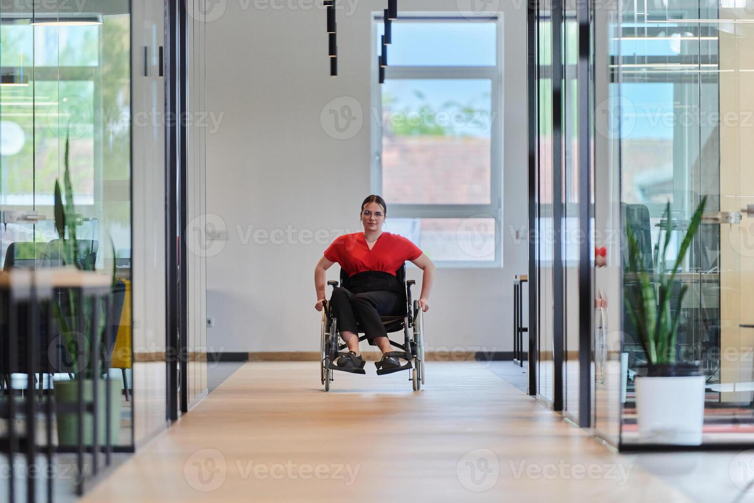 A modern young businesswoman in a wheelchair is surrounded by an inclusive workspace with glass-walled offices, embodying determination and innovation in the business world photo