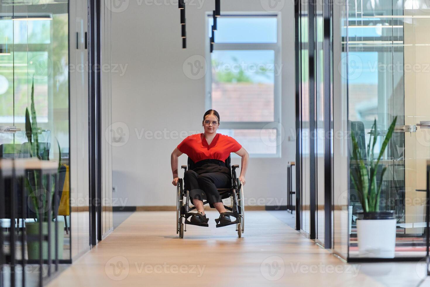 A modern young businesswoman in a wheelchair is surrounded by an inclusive workspace with glass-walled offices, embodying determination and innovation in the business world photo