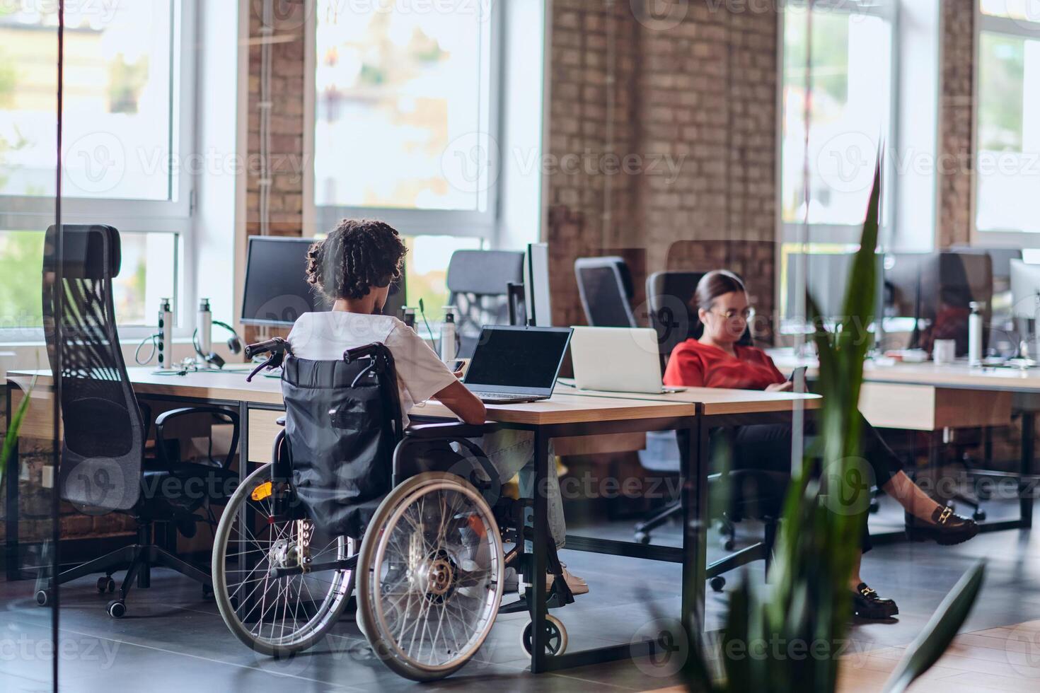 Business colleagues work within a modern startup coworking office, with one of them, an African America in a wheelchair, actively engaging with computers and laptops to collaboratively address various photo
