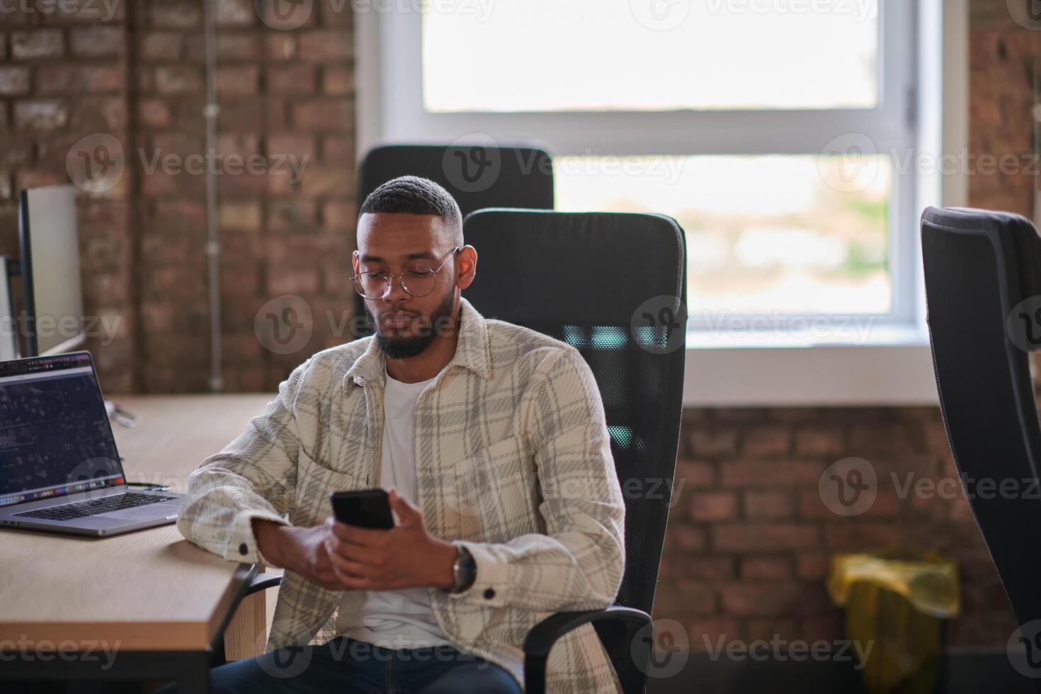 A young African American entrepreneur takes a break in a modern office, using a smartphone to browse social media, capturing a moment of digital connectivity and relaxation amidst his business photo