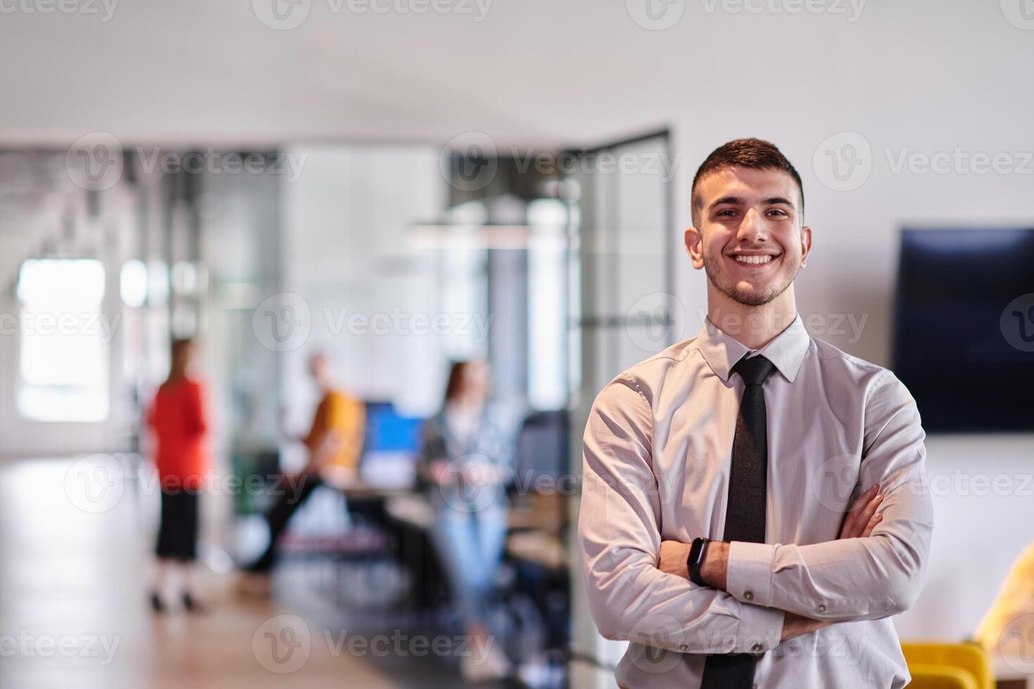 A young business leader stands with crossed arms in a modern office hallway, radiating confidence and a sense of purpose, embodying a dynamic and inspirational presence. photo