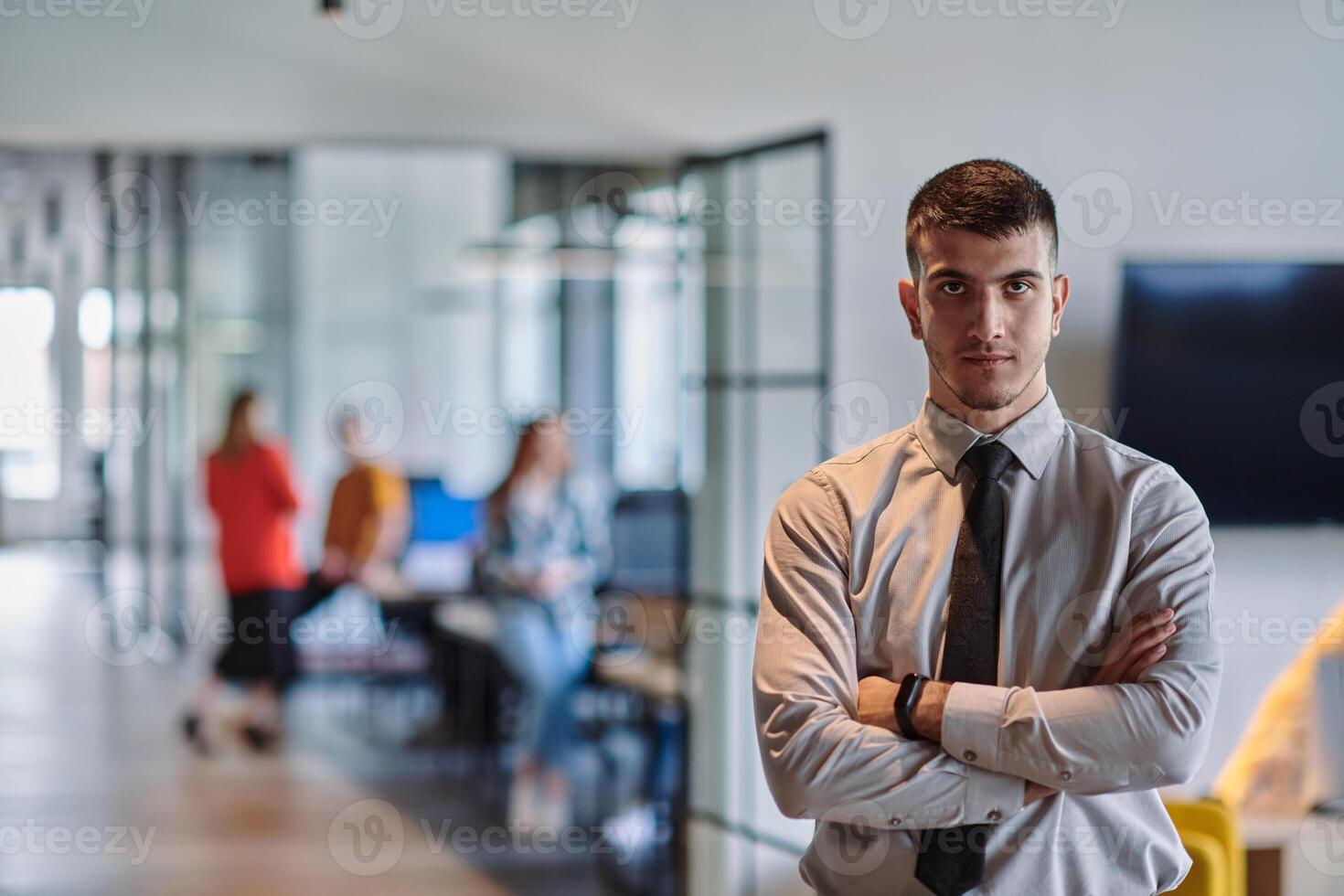 A young business leader stands with crossed arms in a modern office hallway, radiating confidence and a sense of purpose, embodying a dynamic and inspirational presence. photo