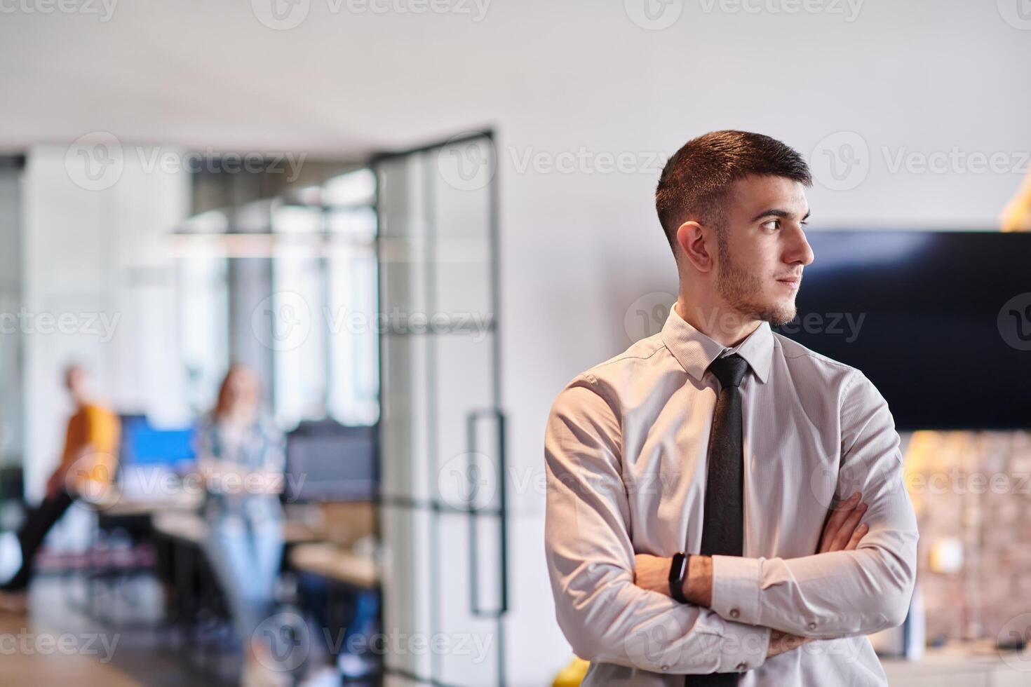 A young business leader stands with crossed arms in a modern office hallway, radiating confidence and a sense of purpose, embodying a dynamic and inspirational presence. photo