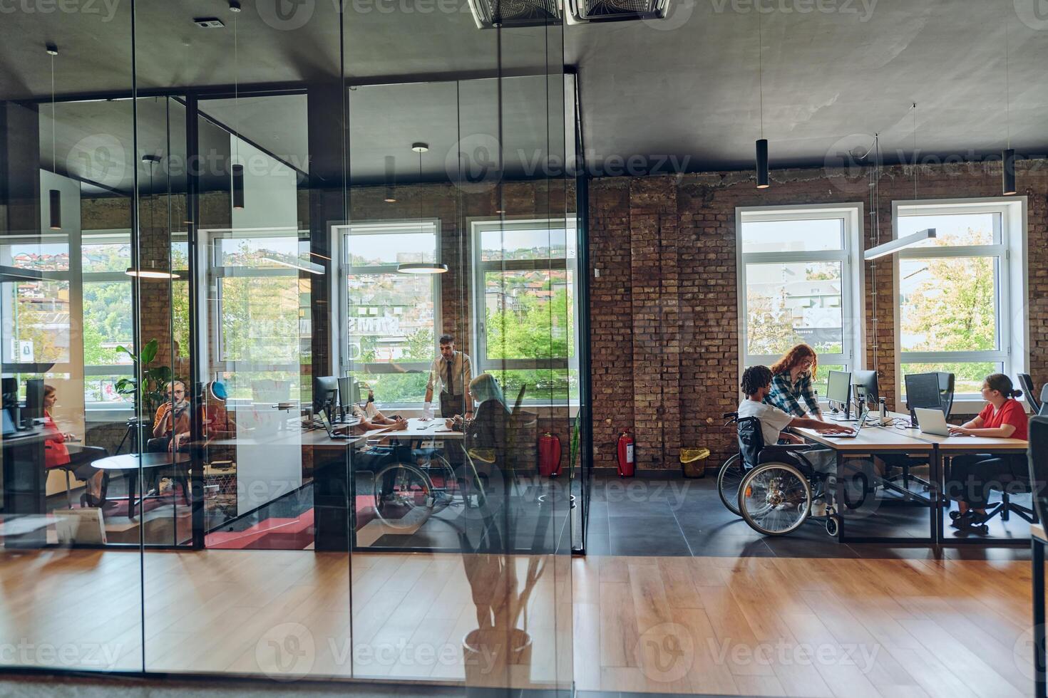 A young business group, including an African American businessman in a wheelchair, collaborates within a modern glass office, actively engaged around a computer and laptop, collectively solving photo