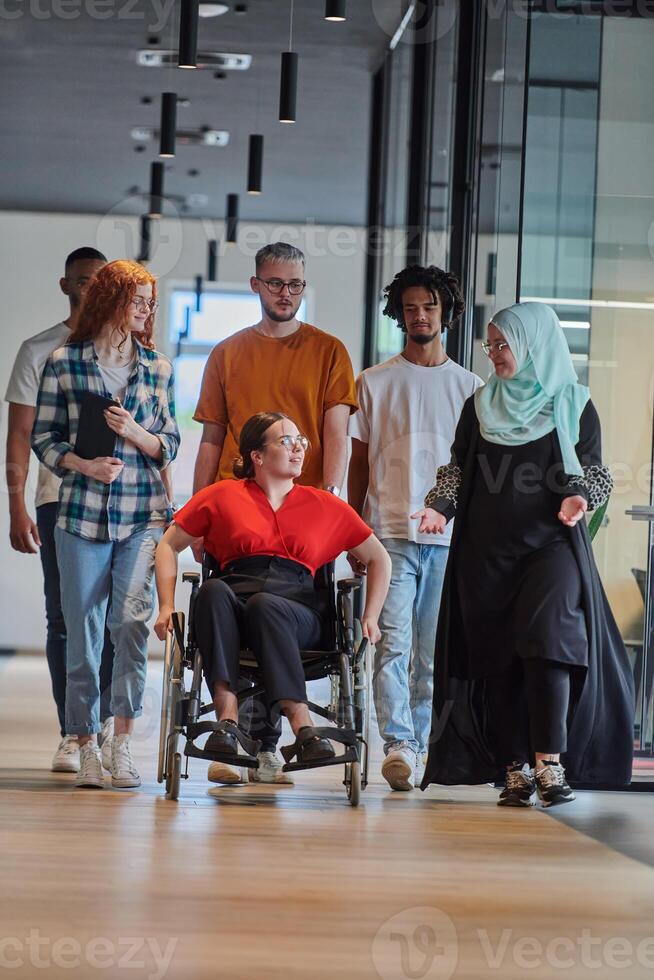 A diverse group of young business people walking a corridor in the glass-enclosed office of a modern startup, including a person in a wheelchair and a woman wearing a hijab, showing a dynamic mix of photo
