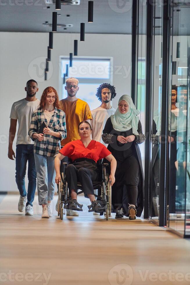 A diverse group of young business people walking a corridor in the glass-enclosed office of a modern startup, including a person in a wheelchair and a woman wearing a hijab, showing a dynamic mix of photo