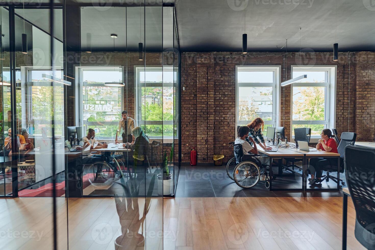 A young business group, including an African American businessman in a wheelchair, collaborates within a modern glass office, actively engaged around a computer and laptop, collectively solving photo