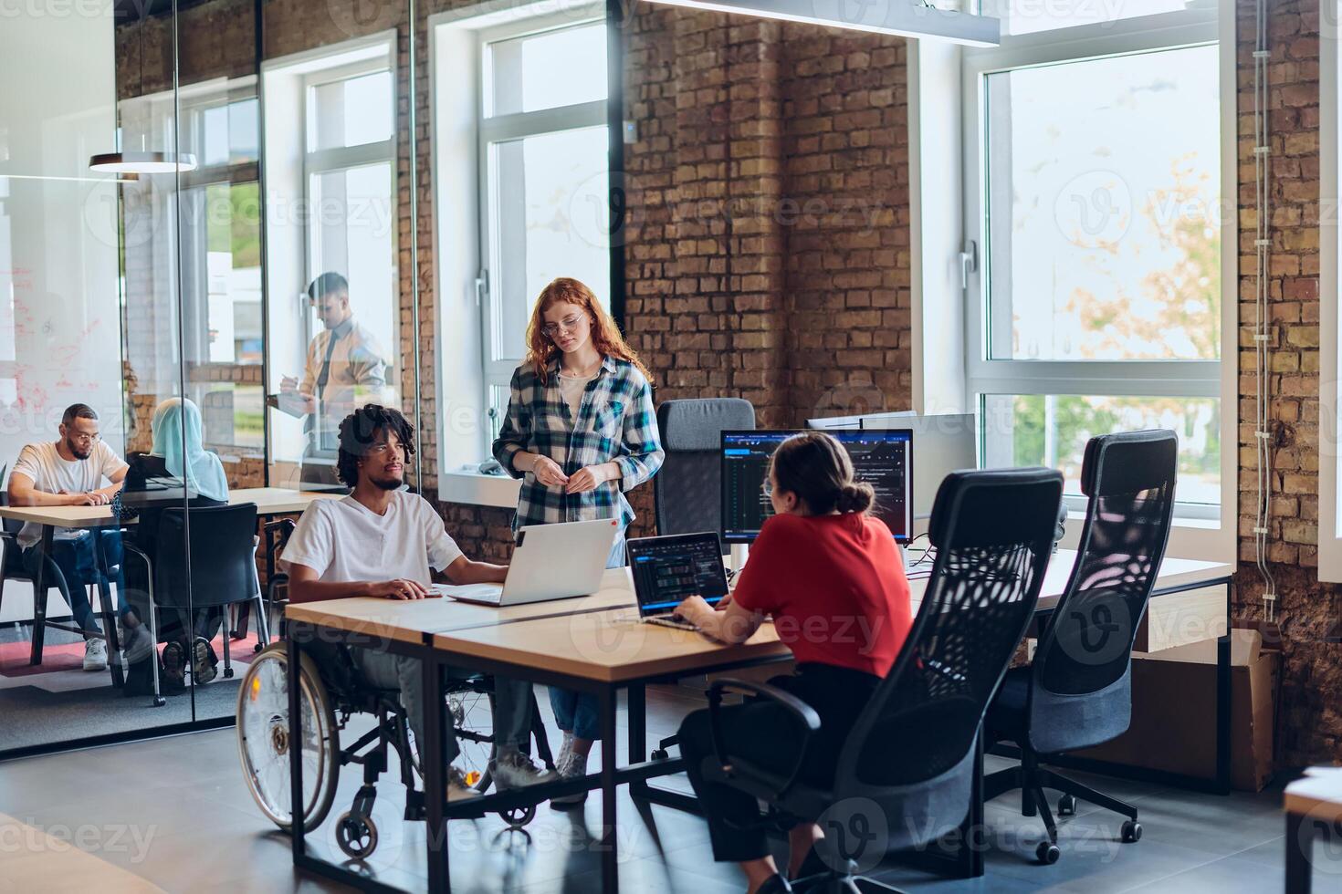 A young business group, including an African American businessman in a wheelchair, collaborates within a modern glass office, actively engaged around a computer and laptop, collectively solving photo