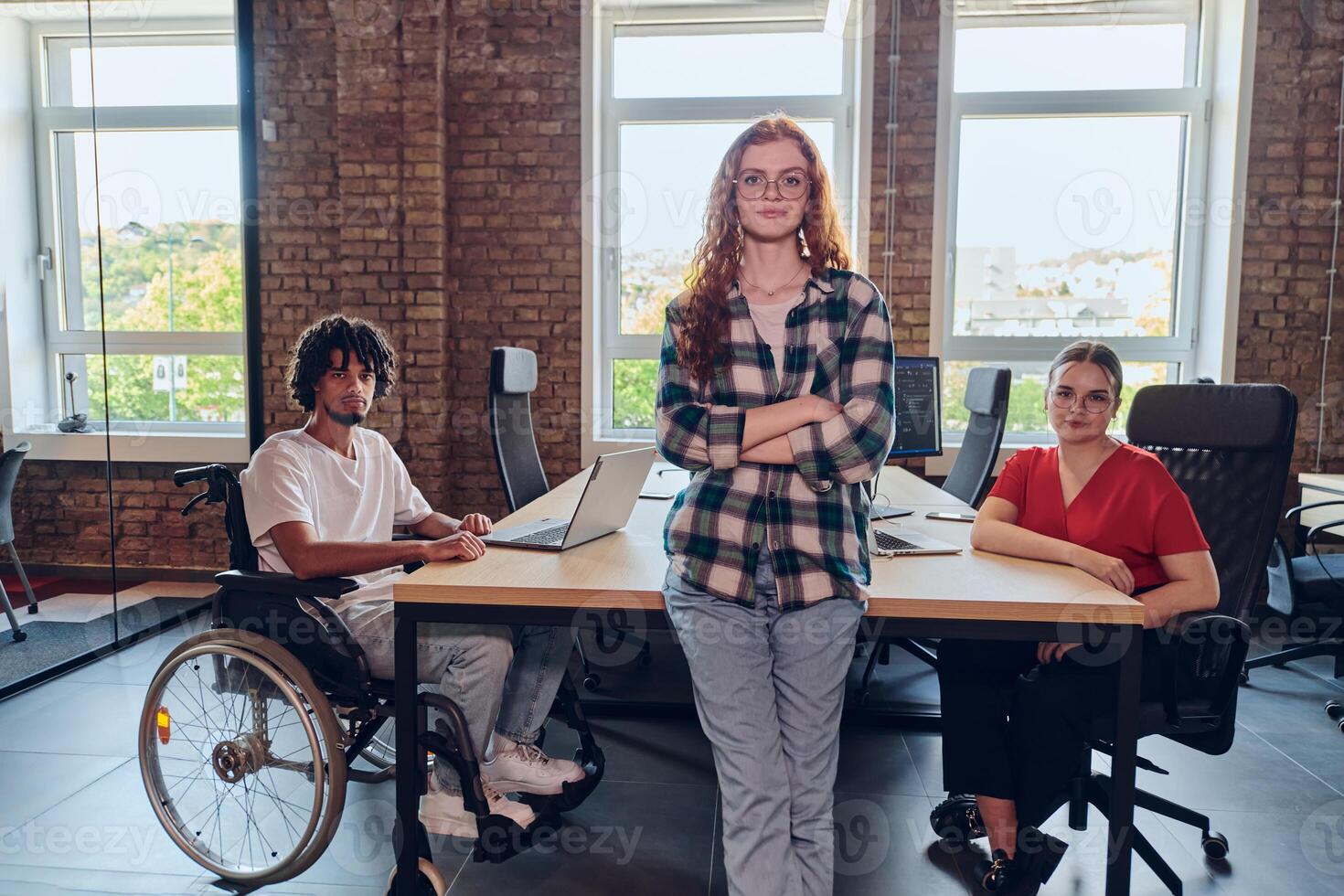A young business group, including an African American businessman in a wheelchair, collaborates within a modern glass office, actively engaged around a computer and laptop, collectively solving photo