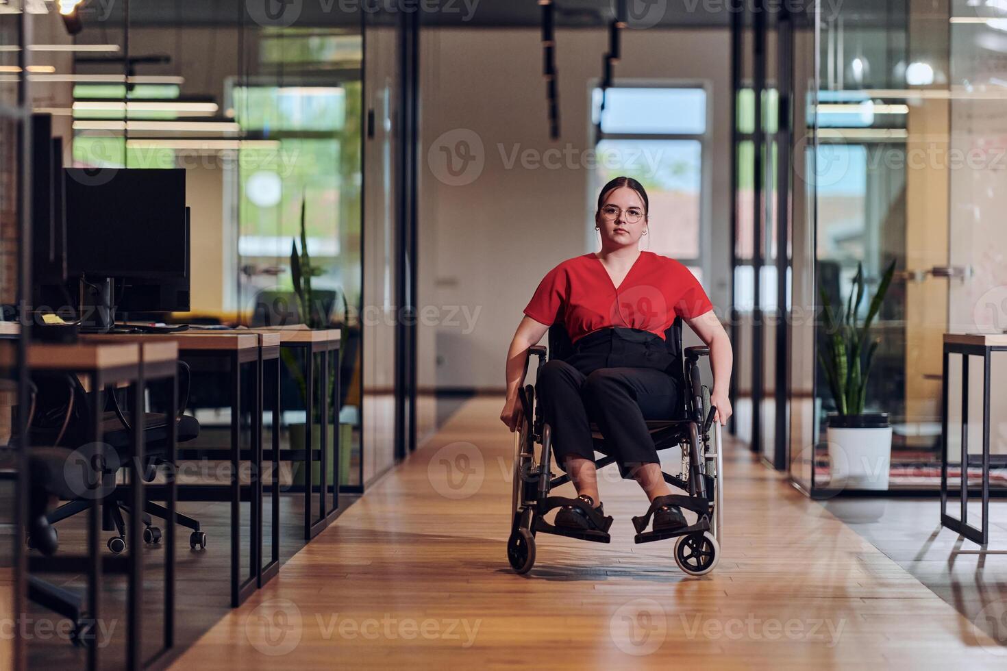 A modern young businesswoman in a wheelchair is surrounded by an inclusive workspace with glass-walled offices, embodying determination and innovation in the business world photo