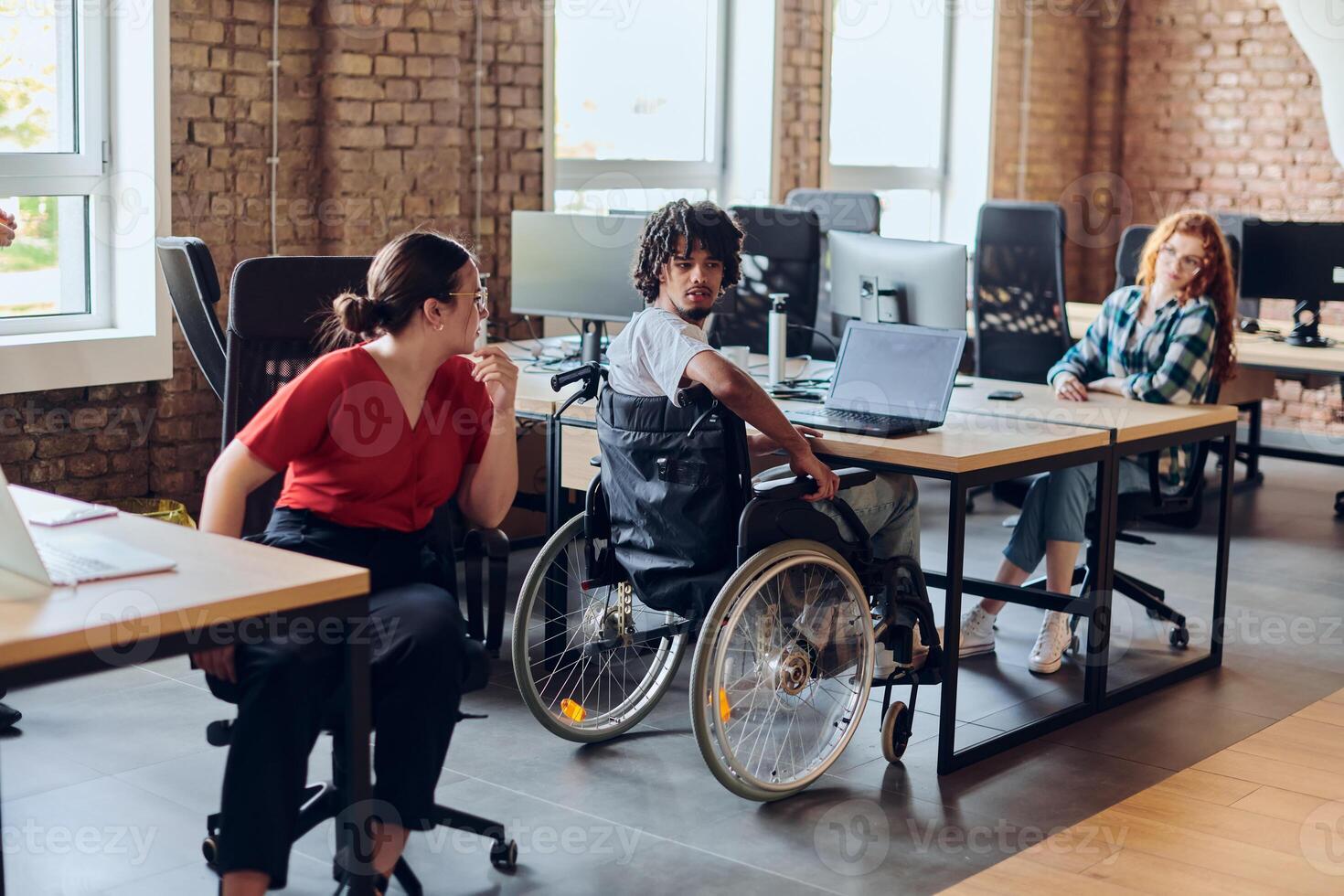 A young business group, including an African American businessman in a wheelchair, collaborates within a modern glass office, actively engaged around a computer and laptop, collectively solving photo