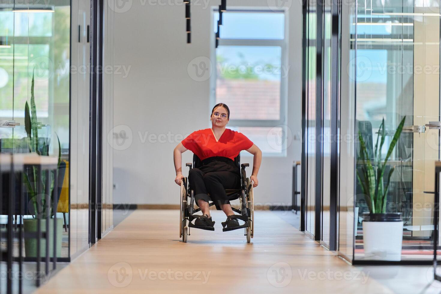 A modern young businesswoman in a wheelchair is surrounded by an inclusive workspace with glass-walled offices, embodying determination and innovation in the business world photo