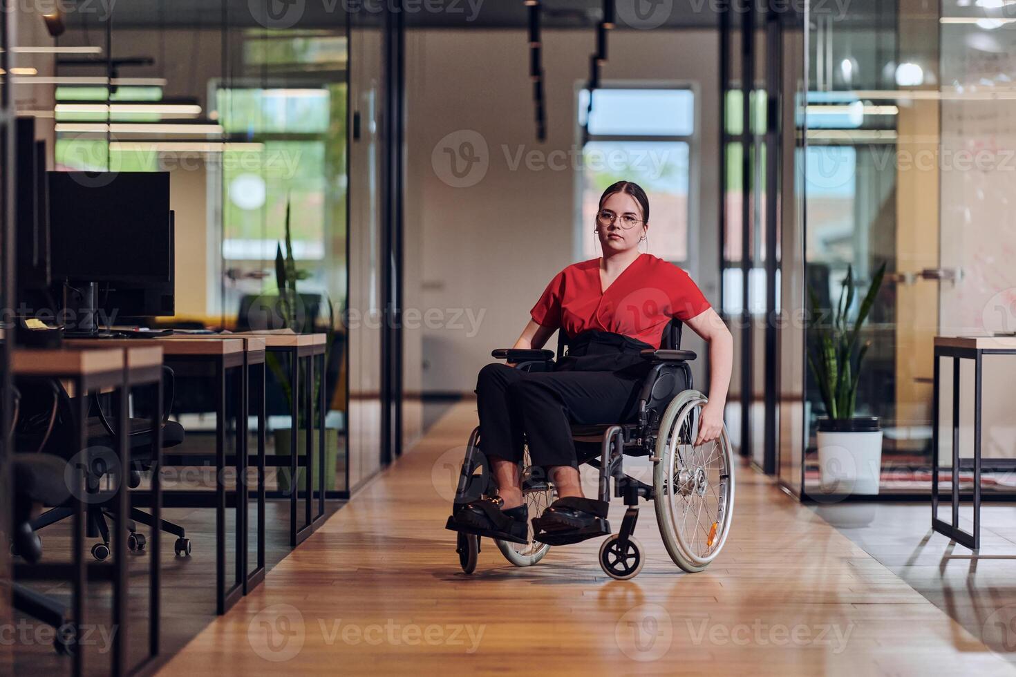A modern young businesswoman in a wheelchair is surrounded by an inclusive workspace with glass-walled offices, embodying determination and innovation in the business world photo