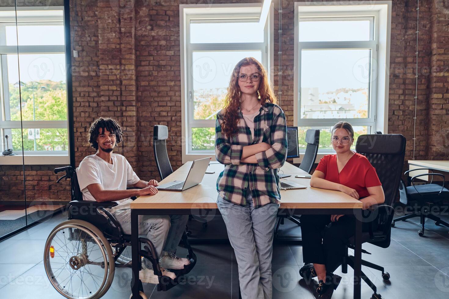 A young business group, including an African American businessman in a wheelchair, collaborates within a modern glass office, actively engaged around a computer and laptop, collectively solving photo