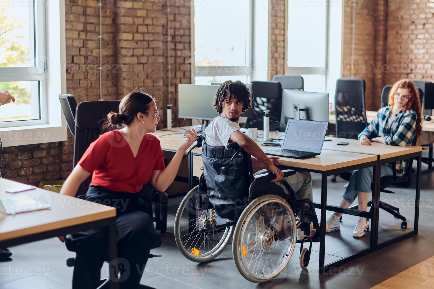 A young business group, including an African American businessman in a wheelchair, collaborates within a modern glass office, actively engaged around a computer and laptop, collectively solving photo