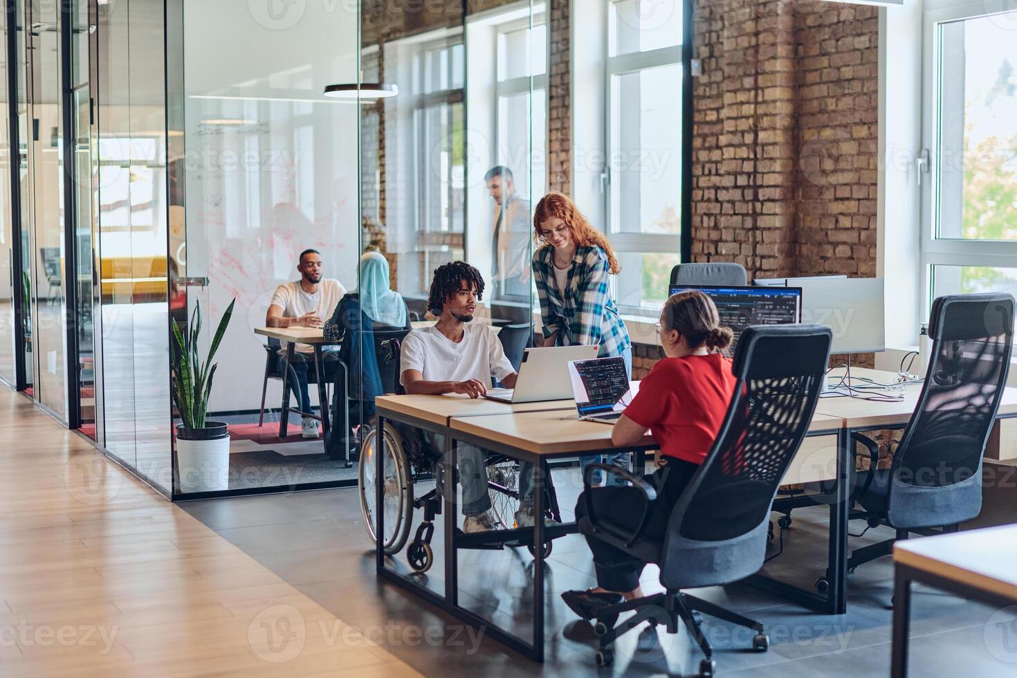 A young business group, including an African American businessman in a wheelchair, collaborates within a modern glass office, actively engaged around a computer and laptop, collectively solving photo