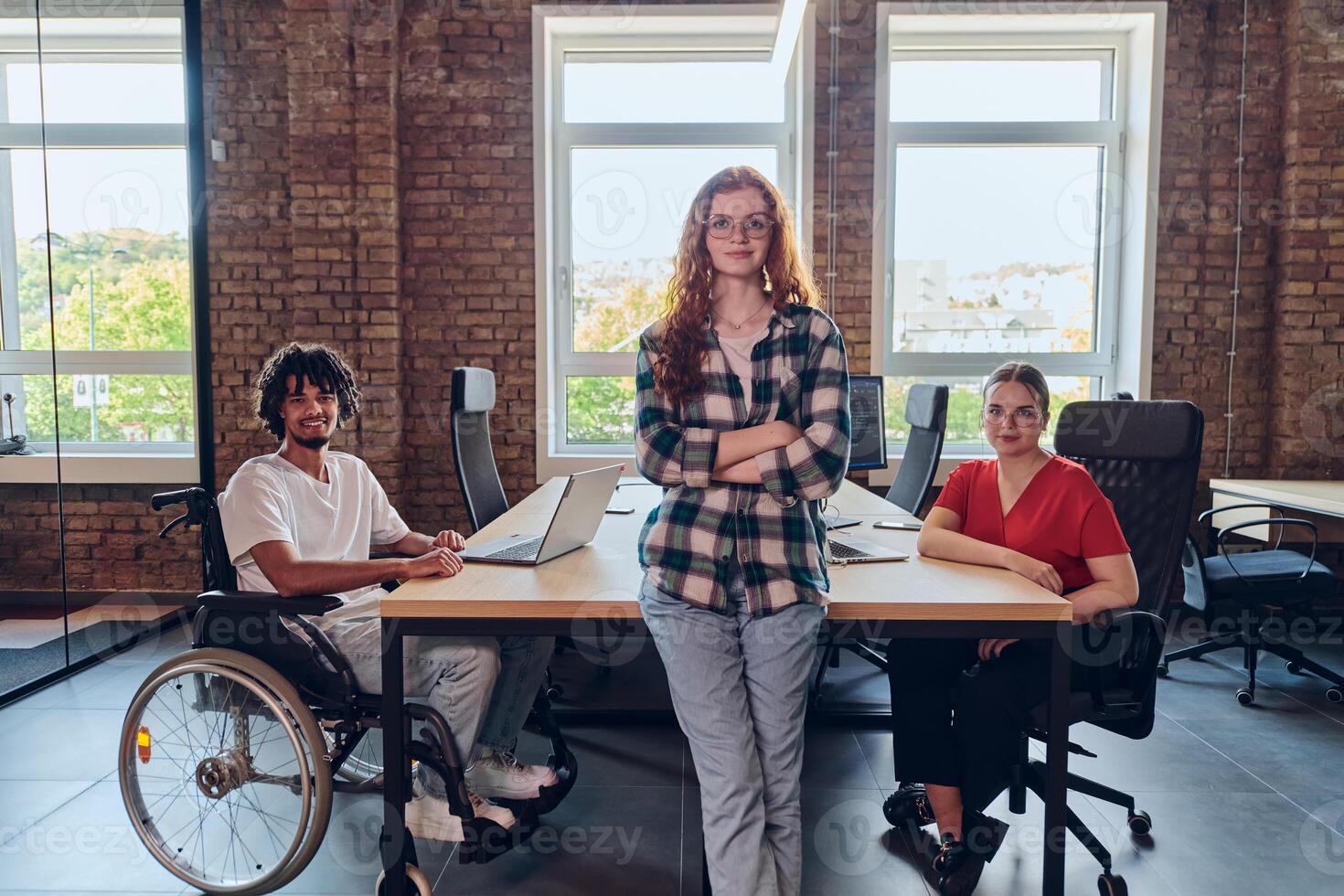 A young business group, including an African American businessman in a wheelchair, collaborates within a modern glass office, actively engaged around a computer and laptop, collectively solving photo