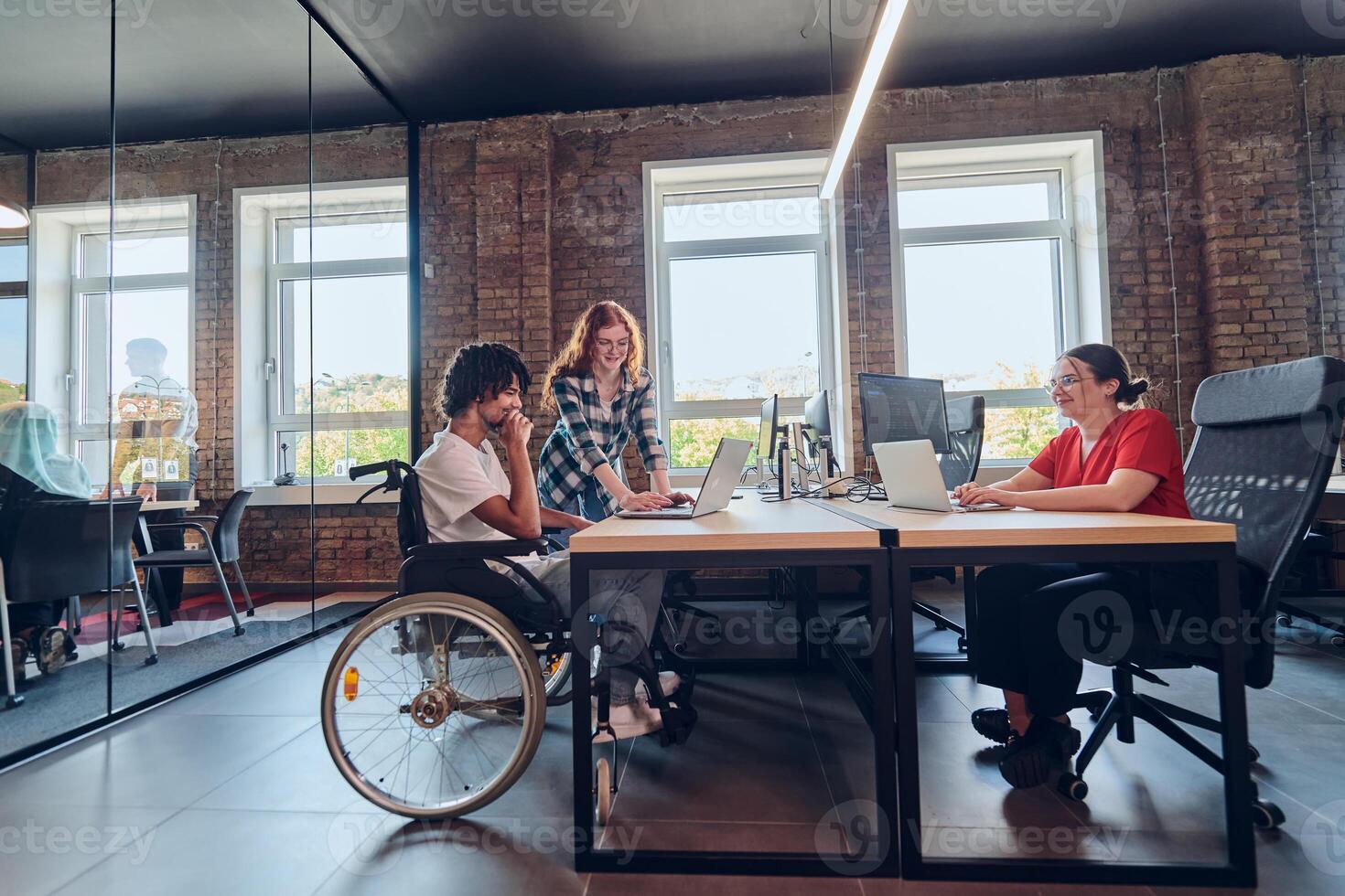 A young business group, including an African American businessman in a wheelchair, collaborates within a modern glass office, actively engaged around a computer and laptop, collectively solving photo