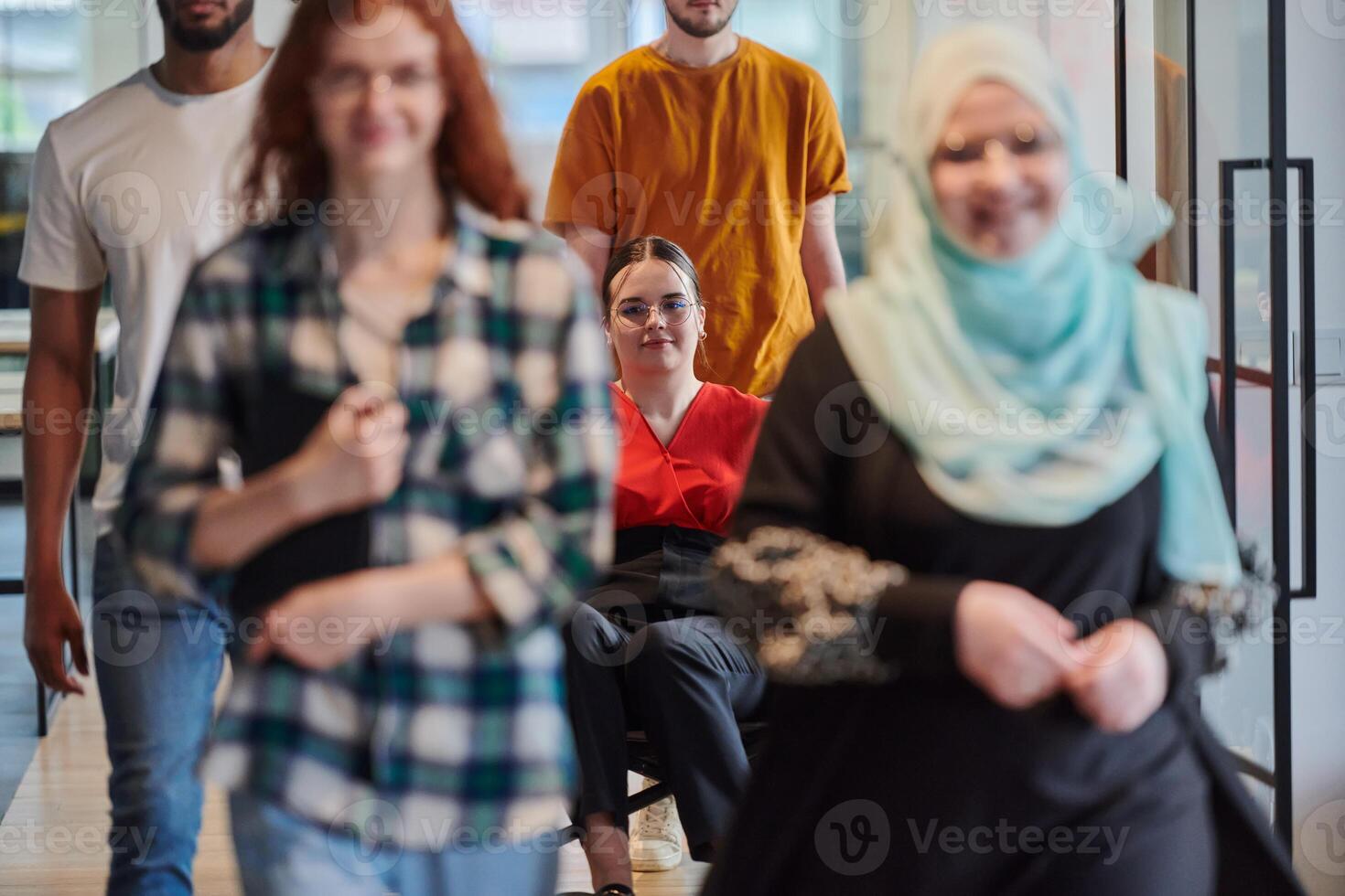A diverse group of business people walking a corridor in the glass-enclosed office of a modern startup, including a person in a wheelchair and a woman wearing a hijab, showing a dynamic mix of photo
