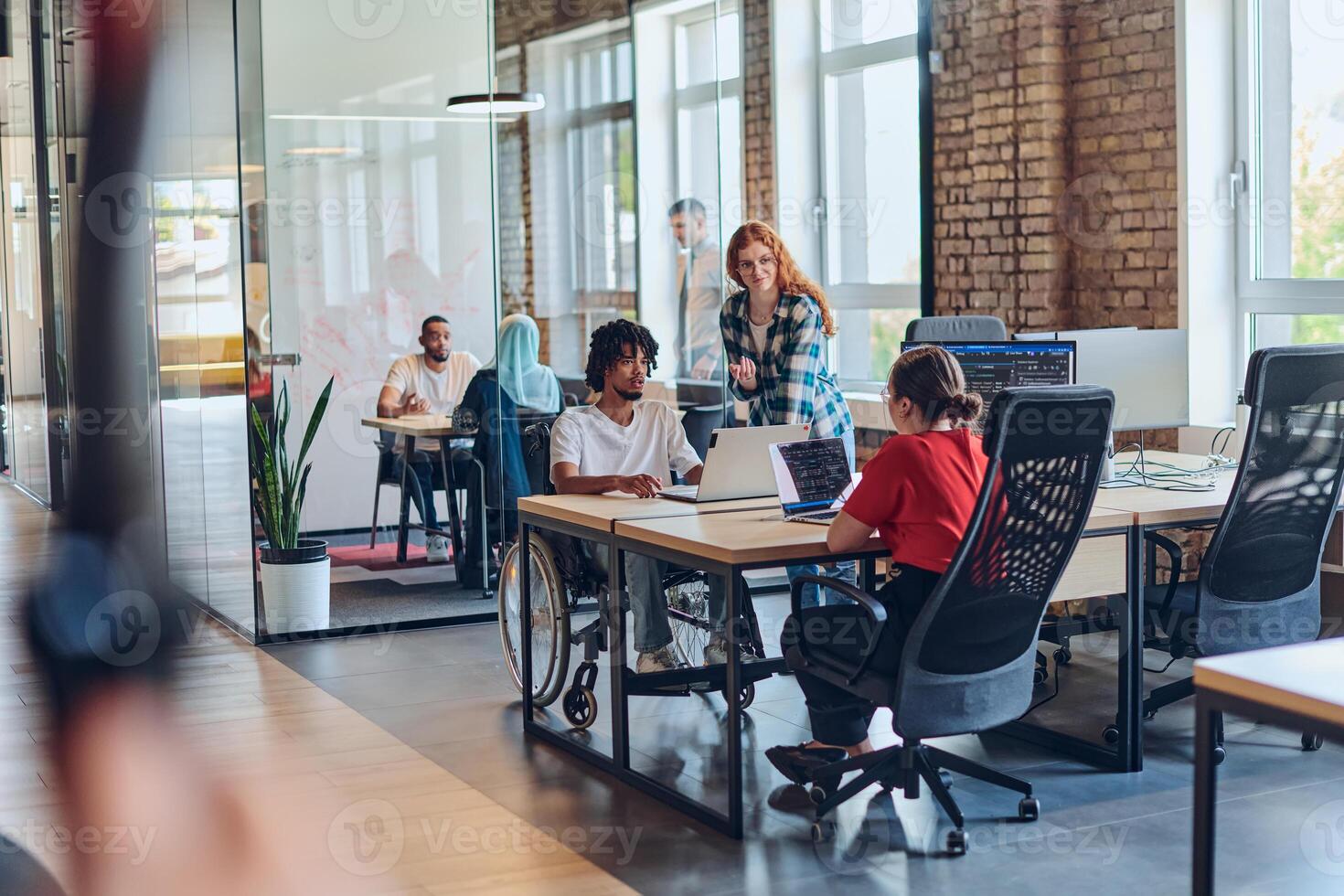 A young business group, including an African American businessman in a wheelchair, collaborates within a modern glass office, actively engaged around a computer and laptop, collectively solving photo