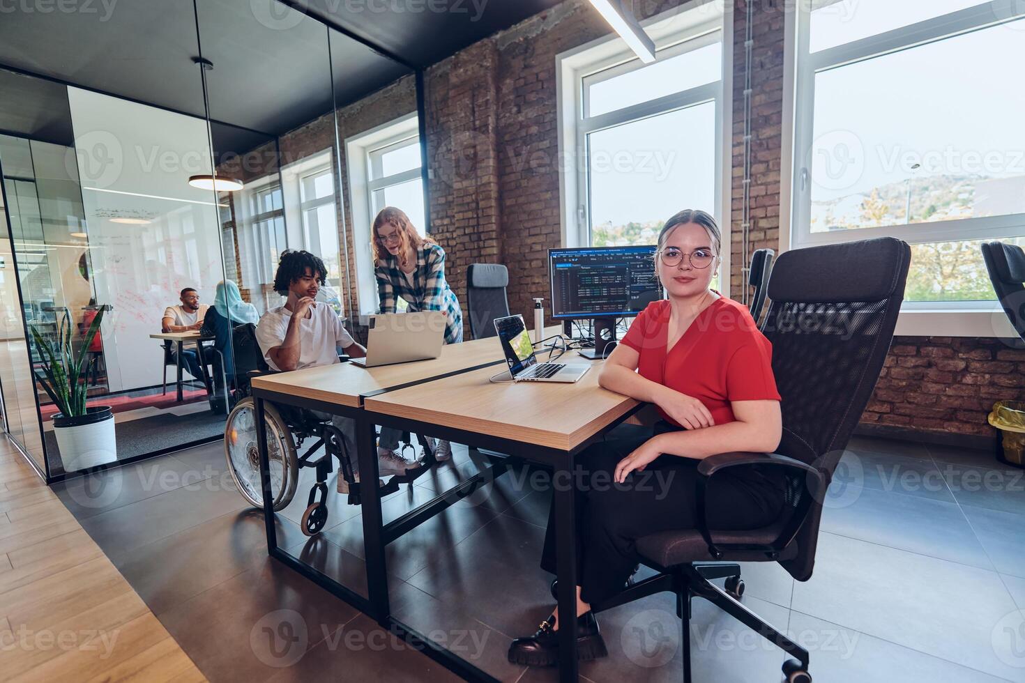 A young business group, including an African American businessman in a wheelchair, collaborates within a modern glass office, actively engaged around a computer and laptop, collectively solving photo