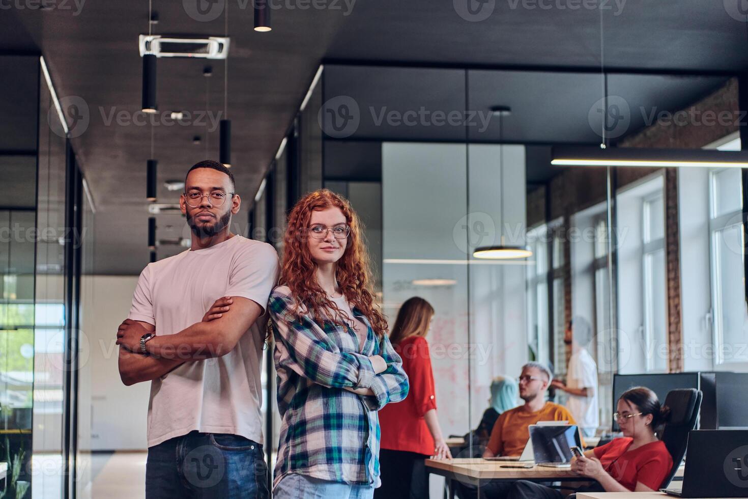A young African American businessman and a modern businesswoman with orange hair stand side by side, arms crossed, exuding confidence and unity in a contemporary office setting, epitomizing dynamic photo