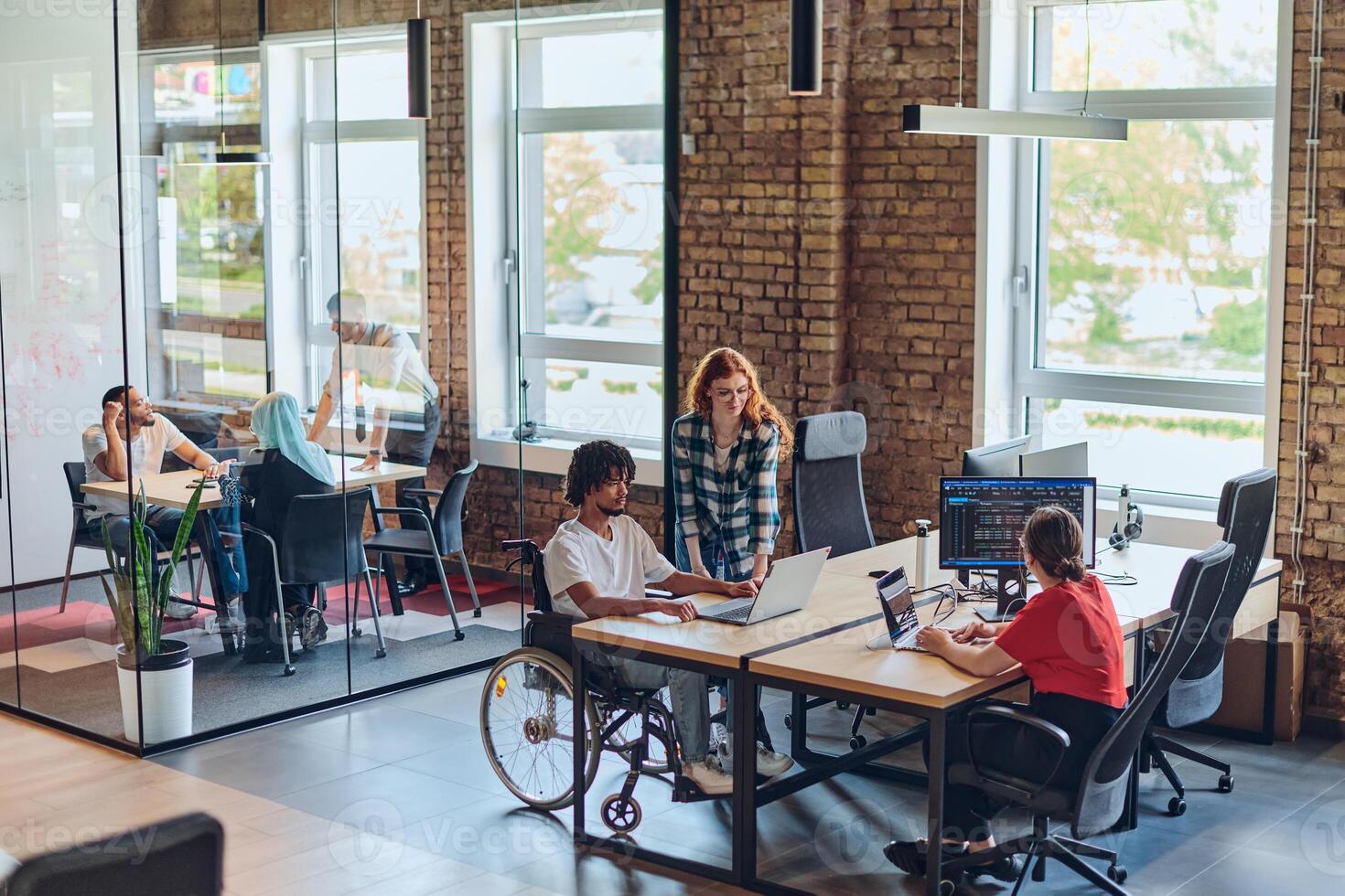 A young business group, including an African American businessman in a wheelchair, collaborates within a modern glass office, actively engaged around a computer and laptop, collectively solving photo