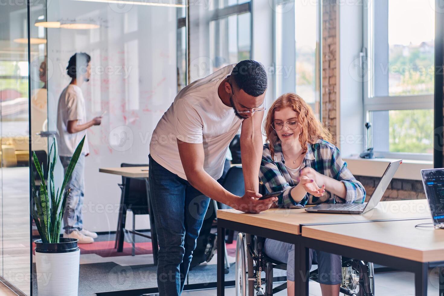 A business group, including an African American businessman in a wheelchair, collaborates within a modern glass office, actively engaged around a computer and laptop, collectively solving diverse photo