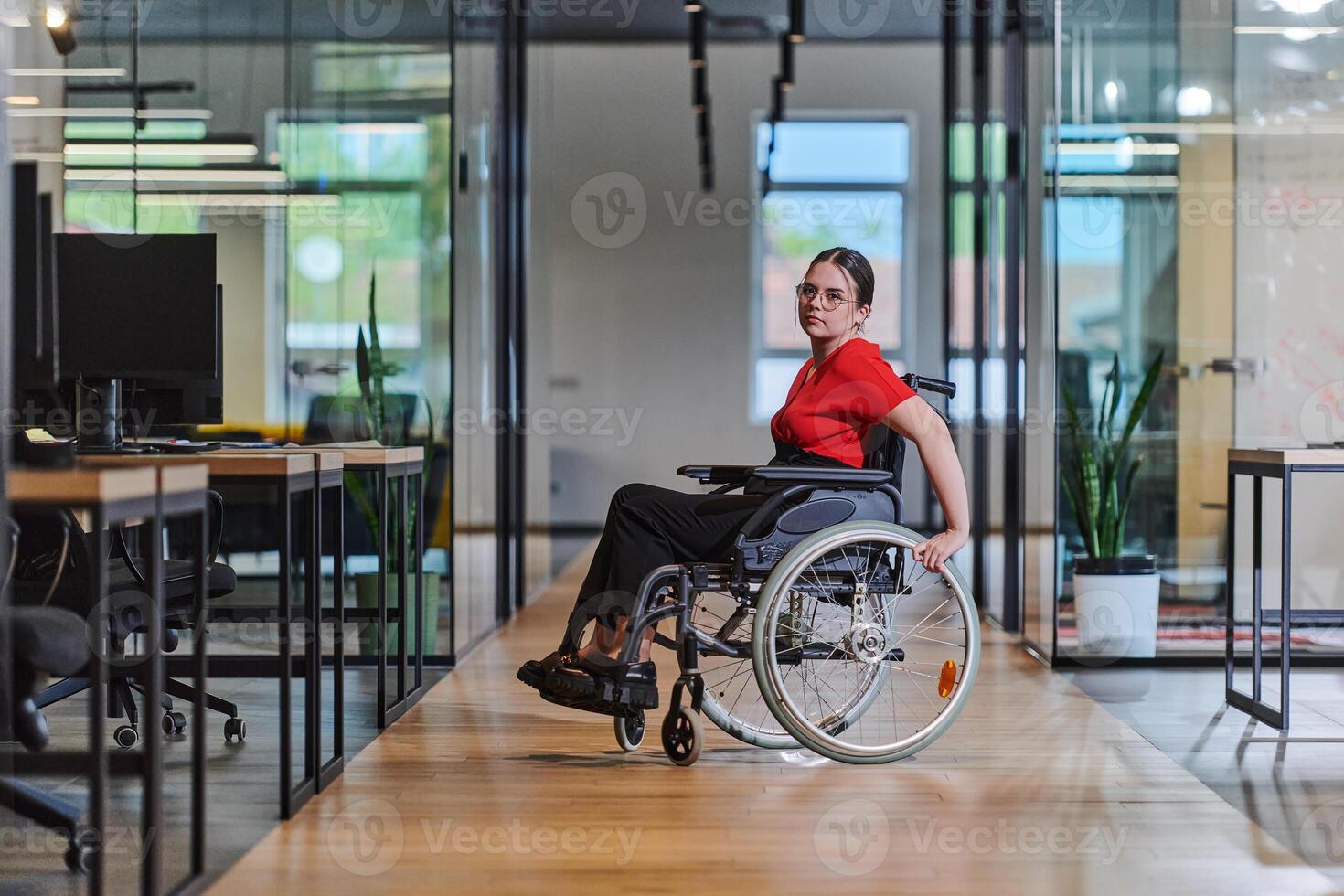 A modern young businesswoman in a wheelchair is surrounded by an inclusive workspace with glass-walled offices, embodying determination and innovation in the business world photo