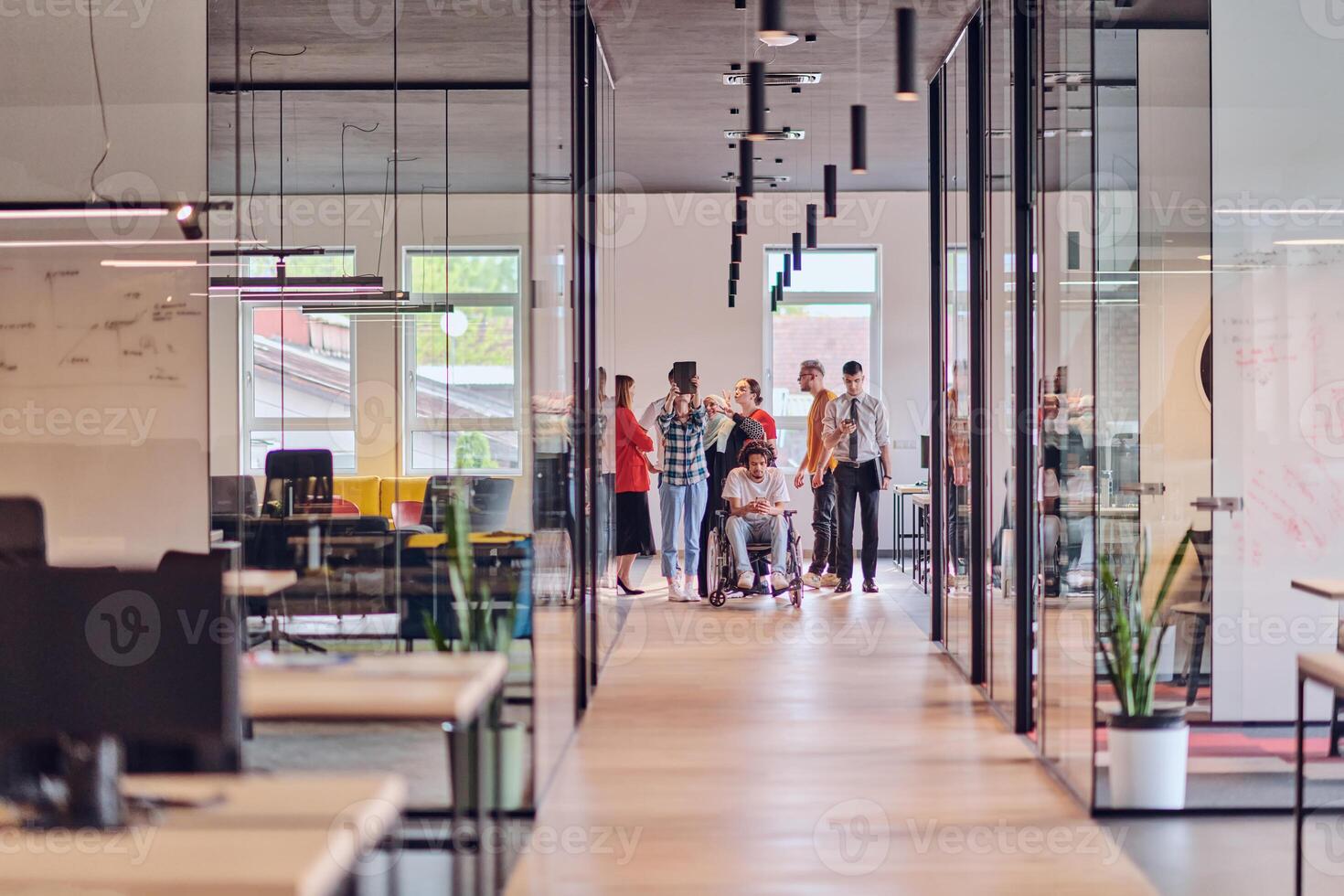 A diverse group of young business people congregates within a modern startup's glass-enclosed office, featuring inclusivity with a person in a wheelchair, an African American young man , and a hijab photo