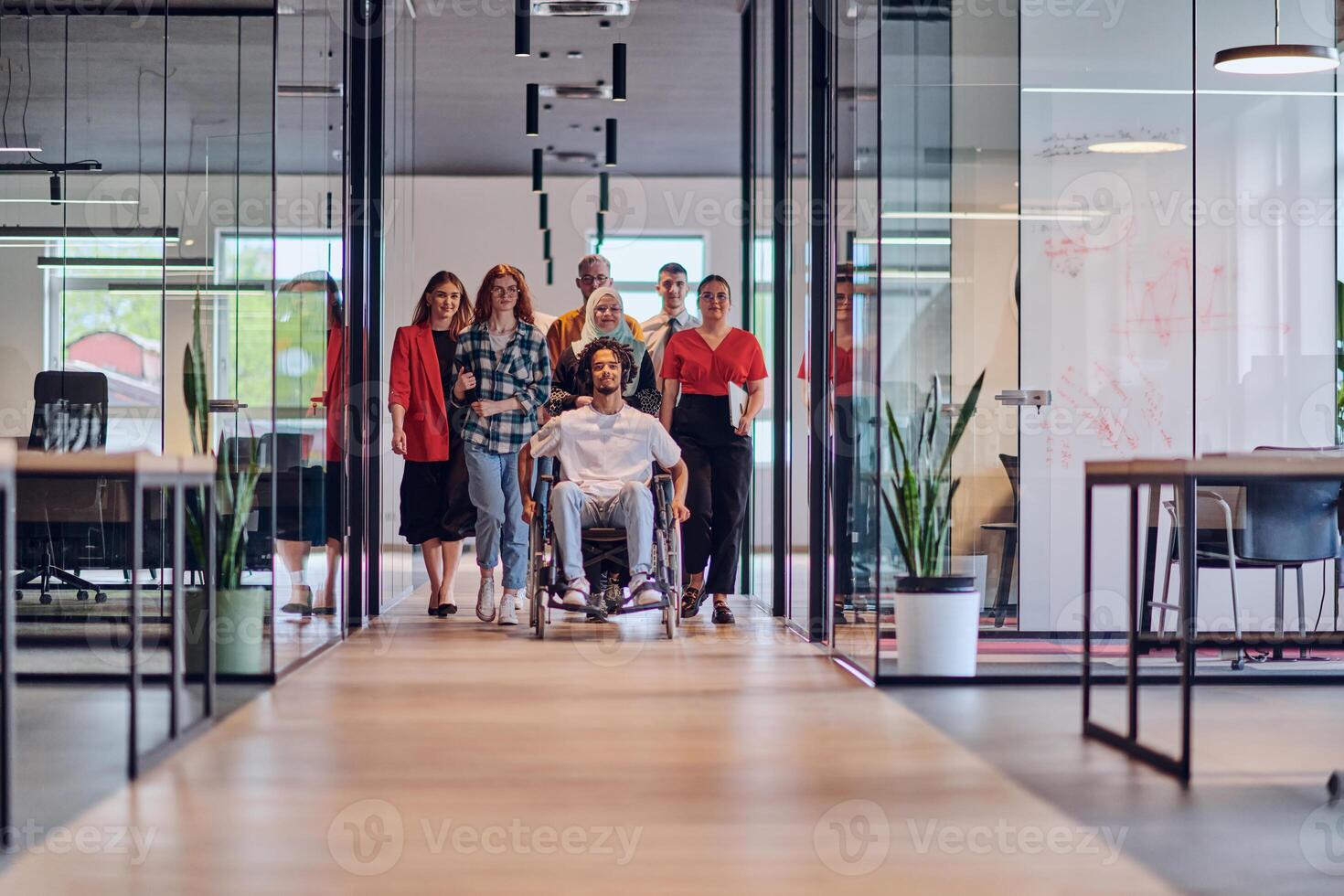 A diverse group of young business people congregates within a modern startup's glass-enclosed office, featuring inclusivity with a person in a wheelchair, an African American young man , and a hijab photo