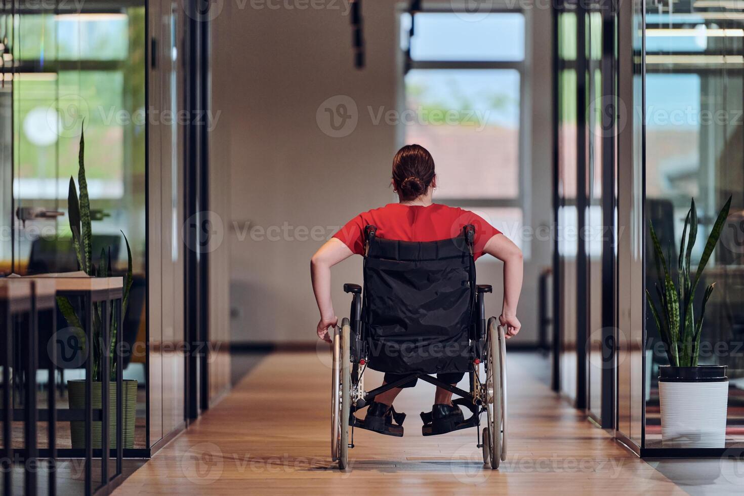 A modern young businesswoman in a wheelchair is surrounded by an inclusive workspace with glass-walled offices, embodying determination and innovation in the business world photo