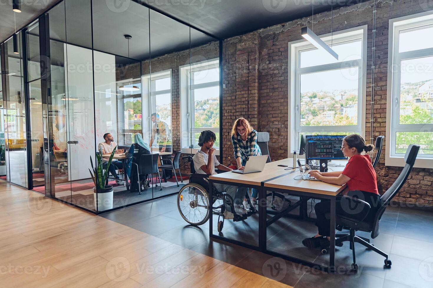A young business group, including an African American businessman in a wheelchair, collaborates within a modern glass office, actively engaged around a computer and laptop, collectively solving photo